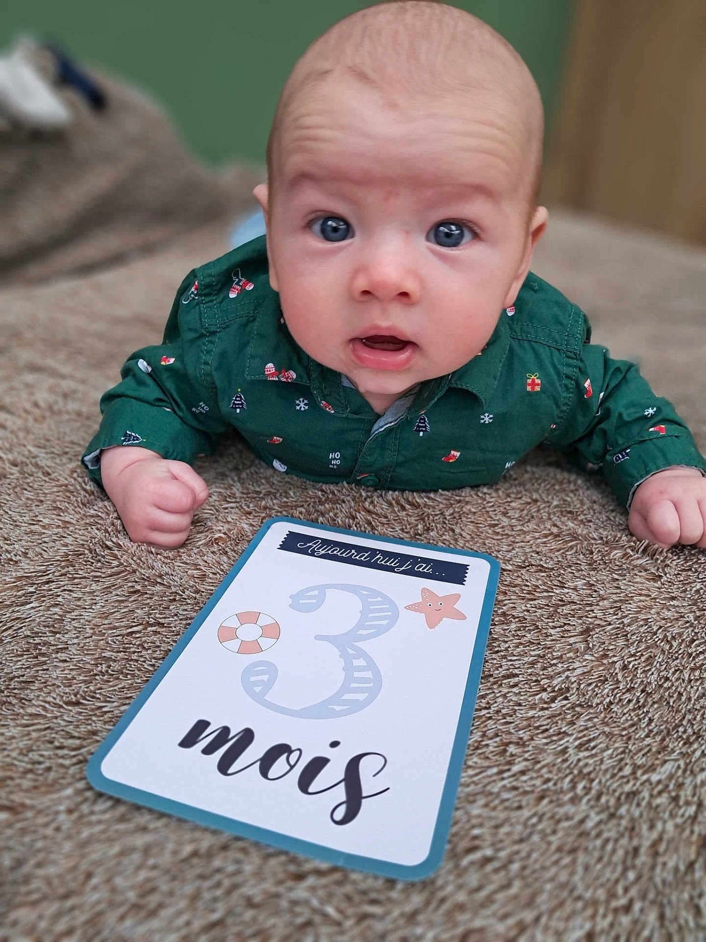 Noah participe au concours pour gagner de l'argent avec cette photo : baby, infant, child, green_shirt, milestone_card, 3_months, brown_blanket, lying_down, wide_eyes, curious, indoor, close_up, face, hands, cute, portrait, soft_texture, decorative_card, expression, celebration