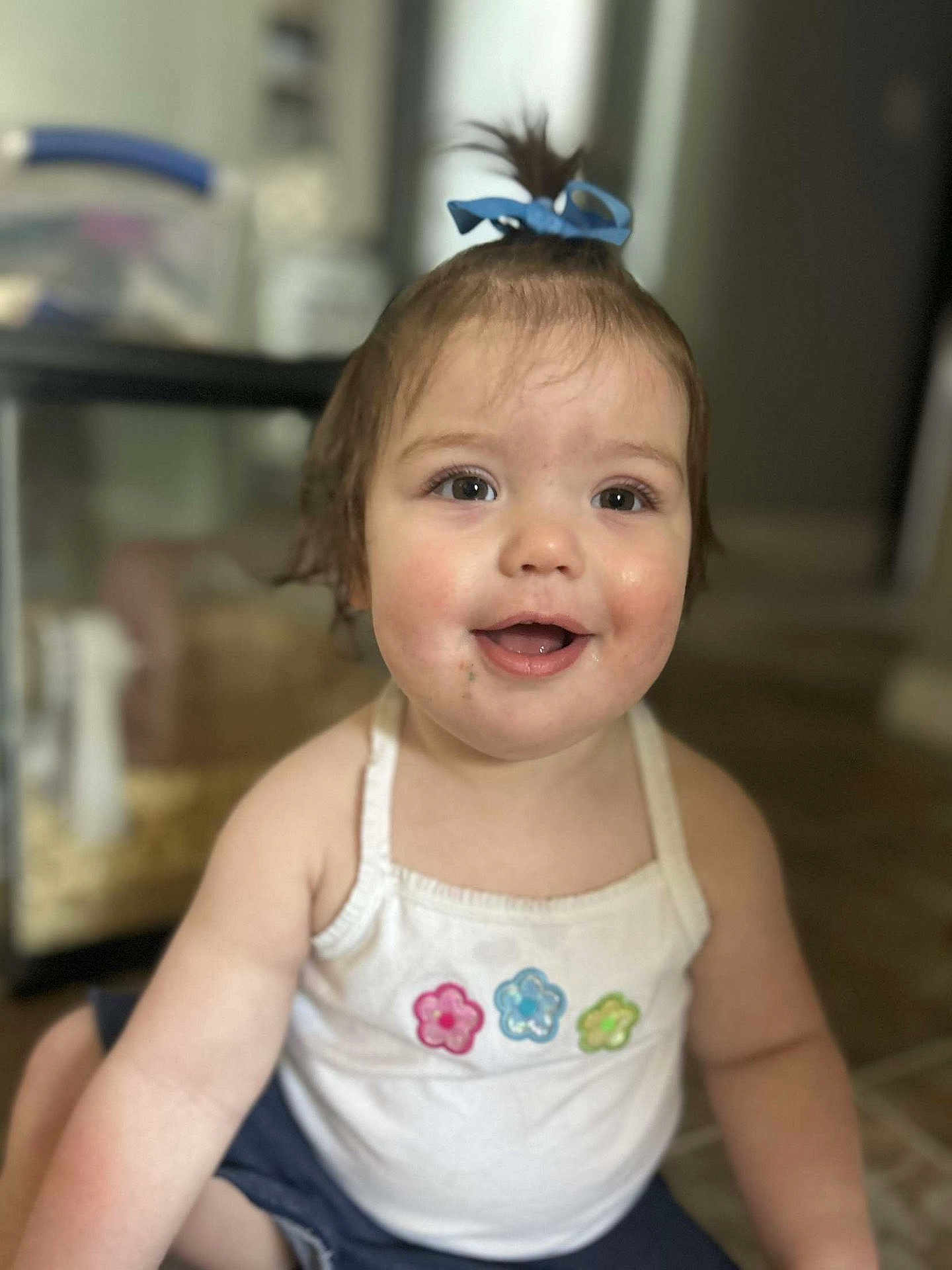 Athena is registered to the contest to win money with this photo: toddler, child, smiling, indoors, tank_top, flower_patch, ribbon, hair_tie, happy, cute, face, skin, eyes, mouth, sitting, floor, background_blur, portrait, person, baby