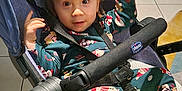 Kassim participe au concours pour gagner de l'argent avec cette photo : toddler, child, stroller, pajamas, indoor, curtains, floor_tiles, furniture, baby, footwear, face, hands, smile, black_stroller, blanket, patterned_clothing, chicco_brand, curly_hair, seated, home