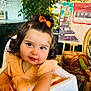 toddler, child, baby, curly_hair, orange_bow, peach_dress, high_chair, indoor, plant, artwork, chair, furniture, curiosity, smile, portrait, cute, young_child, person, face, happy