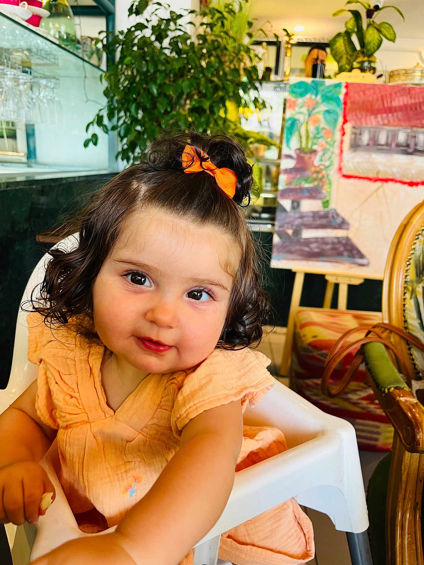 Jalila participe au concours pour gagner de l'argent avec cette photo : toddler, child, baby, curly_hair, orange_bow, peach_dress, high_chair, indoor, plant, artwork, chair, furniture, curiosity, smile, portrait, cute, young_child, person, face, happy