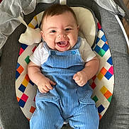 Theo is registered to the contest to win money with this photo: baby, smiling, blue_overalls, infant, baby_seat, colorful_pattern, happy, face, child, cute, clothing, indoor, person, sitting, portrait, young_child, joyful, overalls, fabric, cheerful
