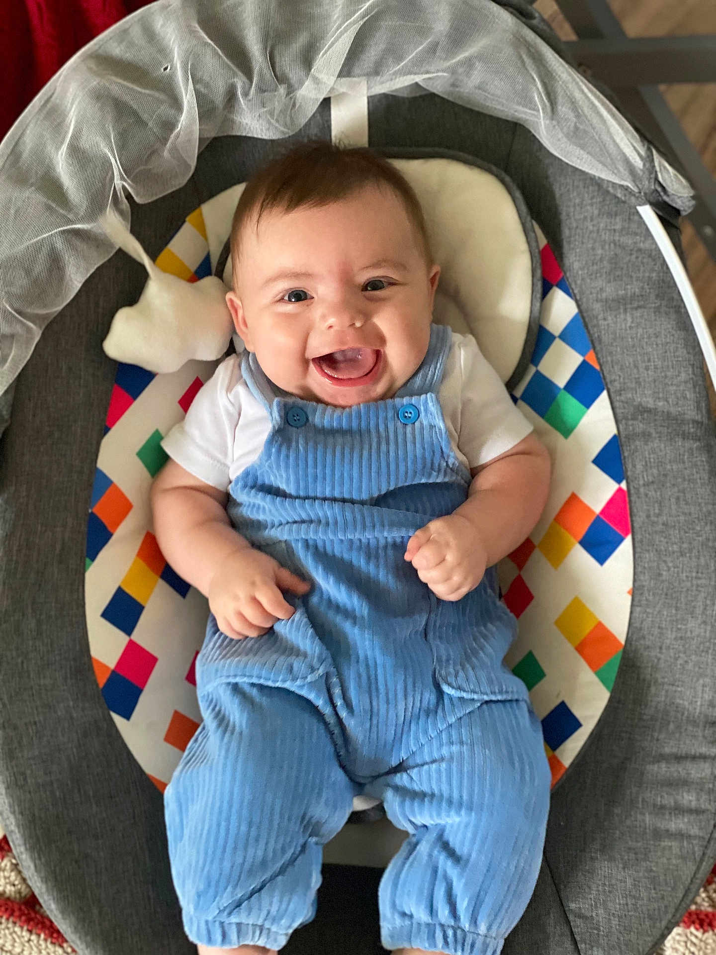 Theo is registered to the contest to win money with this photo: baby, smiling, blue_overalls, infant, baby_seat, colorful_pattern, happy, face, child, cute, clothing, indoor, person, sitting, portrait, young_child, joyful, overalls, fabric, cheerful
