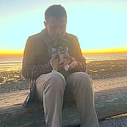 Clyde is registered to the contest to win money with this photo: person, dog, beach, sunset, log, scarf, sand, water, sky, nature, outdoor, casual_clothing, sitting, pet, calm, warm_light, shoes, relaxation, scenic, evening