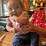 baby, child, infant, pink_bow, pink_clothing, floral_shirt, jeans, socks, table, hand, adult, restaurant, indoor, wooden_table, smiling, cute, person, seated, supporting_hand, chewing_fingers