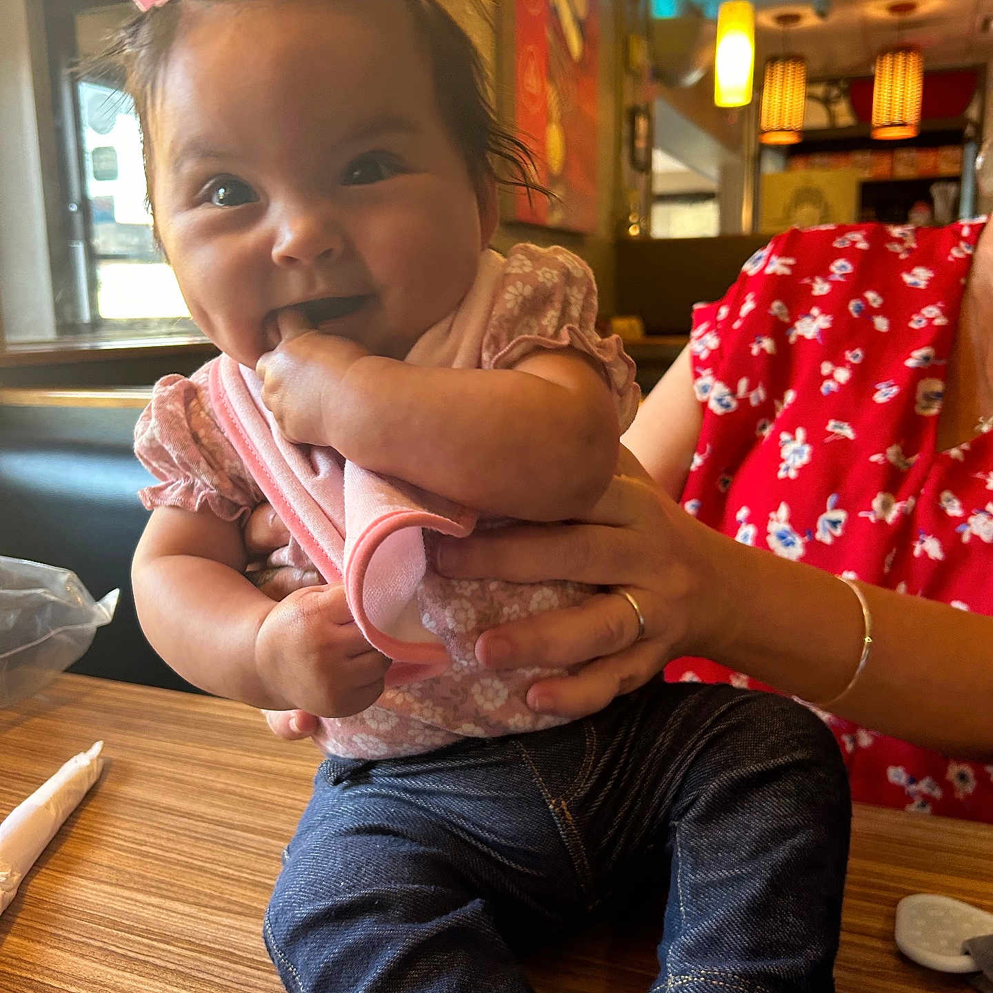 Vada is registered to the contest to win money with this photo: adult, baby, chewing_fingers, child, cute, floral_shirt, hand, indoor, infant, jeans, person, pink_bow, pink_clothing, restaurant, seated, smiling, socks, supporting_hand, table, wooden_table
