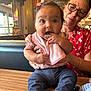 baby, casual_clothing, curious, facial_expression, glasses, holding, indoor, infant, jeans, light_fixture, person, pink_bib, pink_bow, red_flower_shirt, restaurant, seated, supporting_hands, table, woman, wooden_table