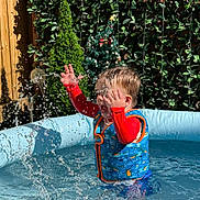 Luca joined the competition — help win amazing prizes! toddler, child, water, splash, pool, life_vest, red_clothing, blue_pool, outdoor, sunny, greenery, plant, fence, summer, play, happy, fun, splashing, water_droplets, daylight