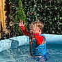 blue_pool, child, daylight, fence, fun, greenery, happy, life_vest, outdoor, plant, play, pool, red_clothing, splash, splashing, summer, sunny, toddler, water, water_droplets