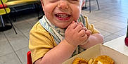Luca joined the competition — help win amazing prizes! toddler, child, smiling, food, chicken_nuggets, french_fries, pacifier, table, restaurant, indoor, yellow_shirt, high_chair, happy, face, hands, casual_dining, background_people, floor_tiles, window, chair