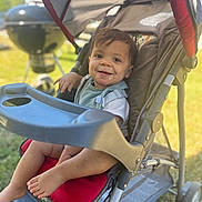 Kashton is registered to the contest to win money with this photo: toddler, child, stroller, outdoor, smiling, baby, seat, red, grass, summer, sunlight, face, foot, plastic, metal, playful, happy, casual, daytime, park