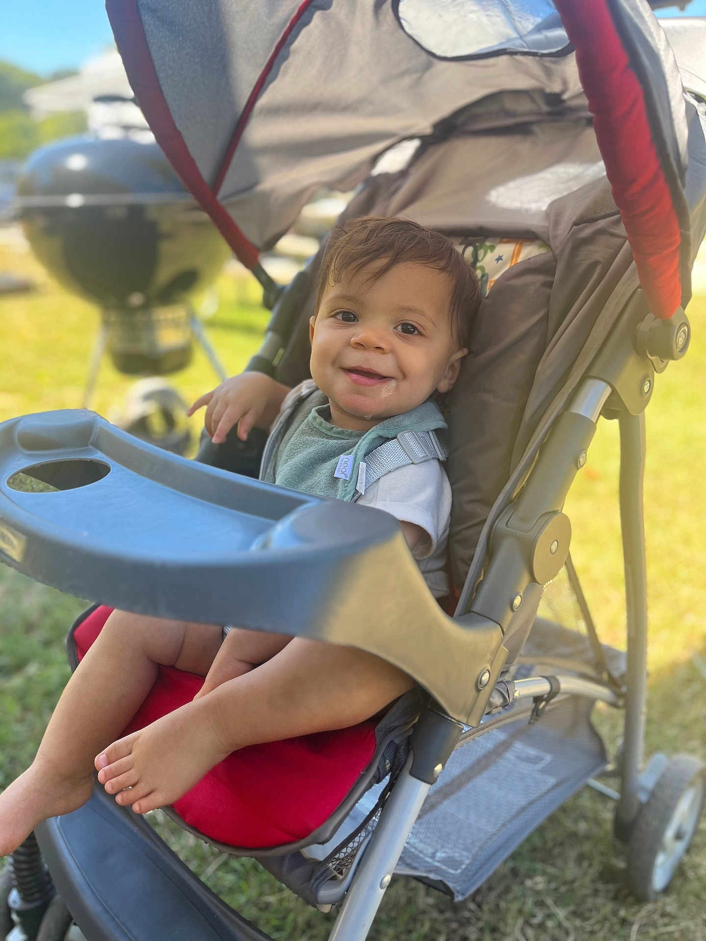 Kashton is registered to the contest to win money with this photo: toddler, child, stroller, outdoor, smiling, baby, seat, red, grass, summer, sunlight, face, foot, plastic, metal, playful, happy, casual, daytime, park