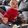 baby, child, infant, smiling, red_sweater, bouncer, baby_seat, living_room, sofa, cushion, cozy, happy, portrait, indoor, hand, sock, footwear, seat_belt, natural_light, cute