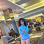 child, curly_hair, blue_shirt, pink_shorts, standing, arms_crossed, indoor, lounge, modern_interior, chairs, table, carpet, ceiling_lights, people, bar_area, mirror, flowers, smile, casual_clothing, relaxed