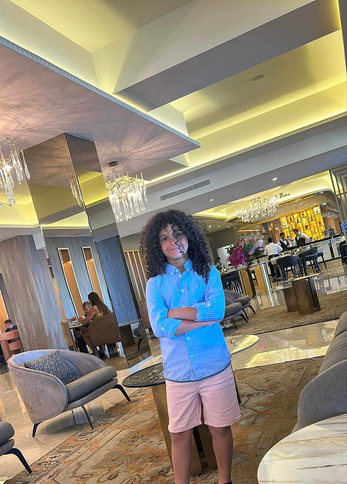 Daniel a rejoint le concours — aidez-le/la à gagner de superbes lots ! child, curly_hair, blue_shirt, pink_shorts, standing, arms_crossed, indoor, lounge, modern_interior, chairs, table, carpet, ceiling_lights, people, bar_area, mirror, flowers, smile, casual_clothing, relaxed