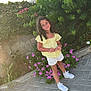 child, girl, smiling, curly_hair, yellow_top, white_shorts, white_shoes, flowers, greenery, outdoor, pavement, plants, sunlight, nature, happy, standing, portrait, casual_clothing, garden, summer