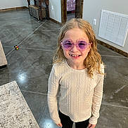 Brenley is registered to the contest to win money with this photo: black_pants, child, concrete_floor, curly_hair, doorway, floor, happy, home, indoor, long_hair, missing_tooth, person, purple_sunglasses, rug, smiling, standing, sunglasses, sweater, toy, wooden_furniture