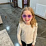 black_pants, child, concrete_floor, curly_hair, doorway, floor, happy, home, indoor, long_hair, missing_tooth, person, purple_sunglasses, rug, smiling, standing, sunglasses, sweater, toy, wooden_furniture