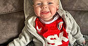 Thomas is registered to the contest to win money with this photo: baby, blond_hair, blue_pants, casual_clothing, child, couch, cozy, cute, face, gray_hoodie, happy, indoor, infant, person, portrait, red_jersey, rug, sitting, smiling, soft_texture