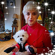 Mookie is registered to the contest to win money with this photo: woman, dog, pet, tuxedo, costume, indoor, elderly, red_sweater, white_dog, fluffy, smiling, holding_pet, lamp, plant, fireplace, wood_floor, decor, jewelry, sparkles, festive