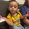 baby, book, brick_wall, casual, child, couch, family, framed_art, gray_pants, happy, indoor, living_room, pittsburgh_steelers, portrait, sibling, sitting, smiling, sports_jersey, wide_eyed, yellow_shirt