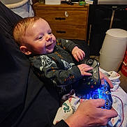 Kingston Jackson joined the competition — help win amazing prizes! baby, infant, smiling, controller, video_game_controller, gaming, adult_hand, blanket, onesie, dresser, bassinet, trash_can, blue_light, hands, couch, portrait, indoors, person, playful, cozy