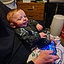 baby, infant, smiling, controller, video_game_controller, gaming, adult_hand, blanket, onesie, dresser, bassinet, trash_can, blue_light, hands, couch, portrait, indoors, person, playful, cozy