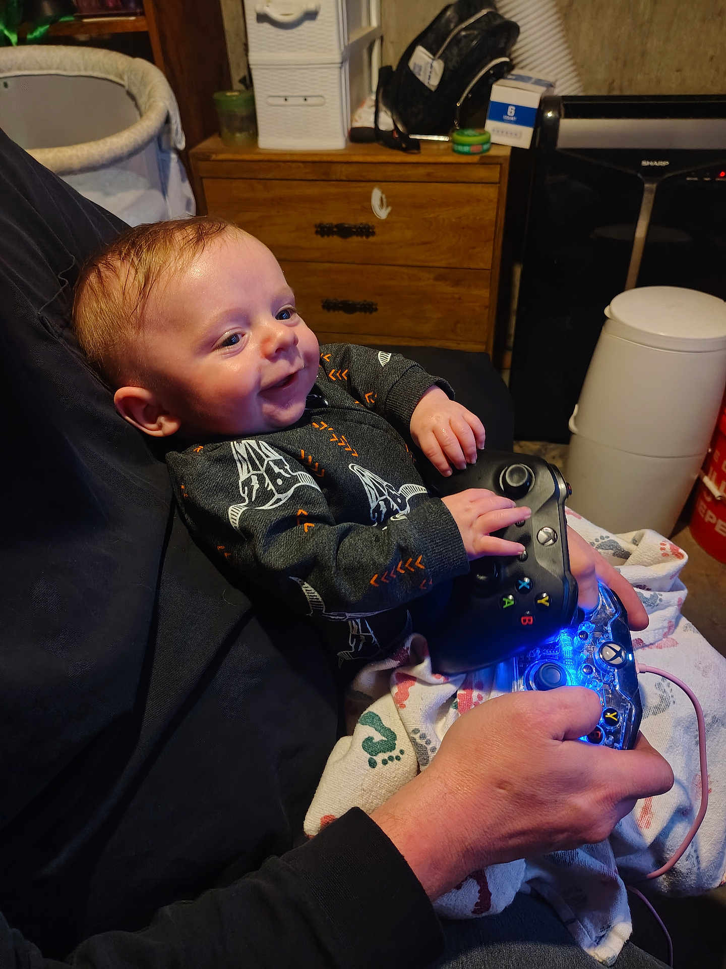 Kingston Jackson joined the competition — help win amazing prizes! baby, infant, smiling, controller, video_game_controller, gaming, adult_hand, blanket, onesie, dresser, bassinet, trash_can, blue_light, hands, couch, portrait, indoors, person, playful, cozy