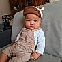 baby, child, cap, overalls, couch, indoor, furniture, wooden_cabinet, living_room, seat, person, portrait, cute, serious, sitting, clothing, infant, face, hands, home