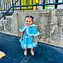 asphalt, blue_dress, cape, child, concrete_wall, cute, daylight, fashion, hair_bows, inflatable, outdoor, person, portrait, railing, shoes, smiling, standing, toddler, urban, yellow_inflatable