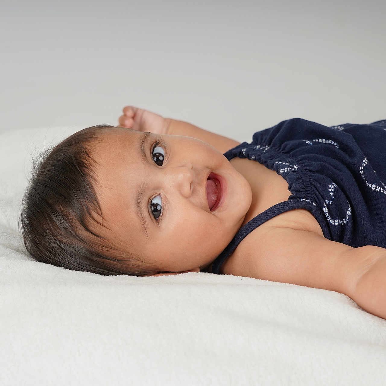 Nikara joined the competition — help win amazing prizes! arms_outstretched, baby, casual_clothing, child, cute, face, happy, head, indoors, infant, looking_at_camera, lying_down, navy_blue_clothing, one_person, portrait, skin, smiling, soft_texture, white_blanket, young_child