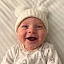 baby, smiling, hat, knit_hat, pom_poms, white_clothing, onesie, infant, happy, face, portrait, lying_down, striped_blanket, soft_texture, cute, child, closeup, indoors, cheerful, wearing_clothes