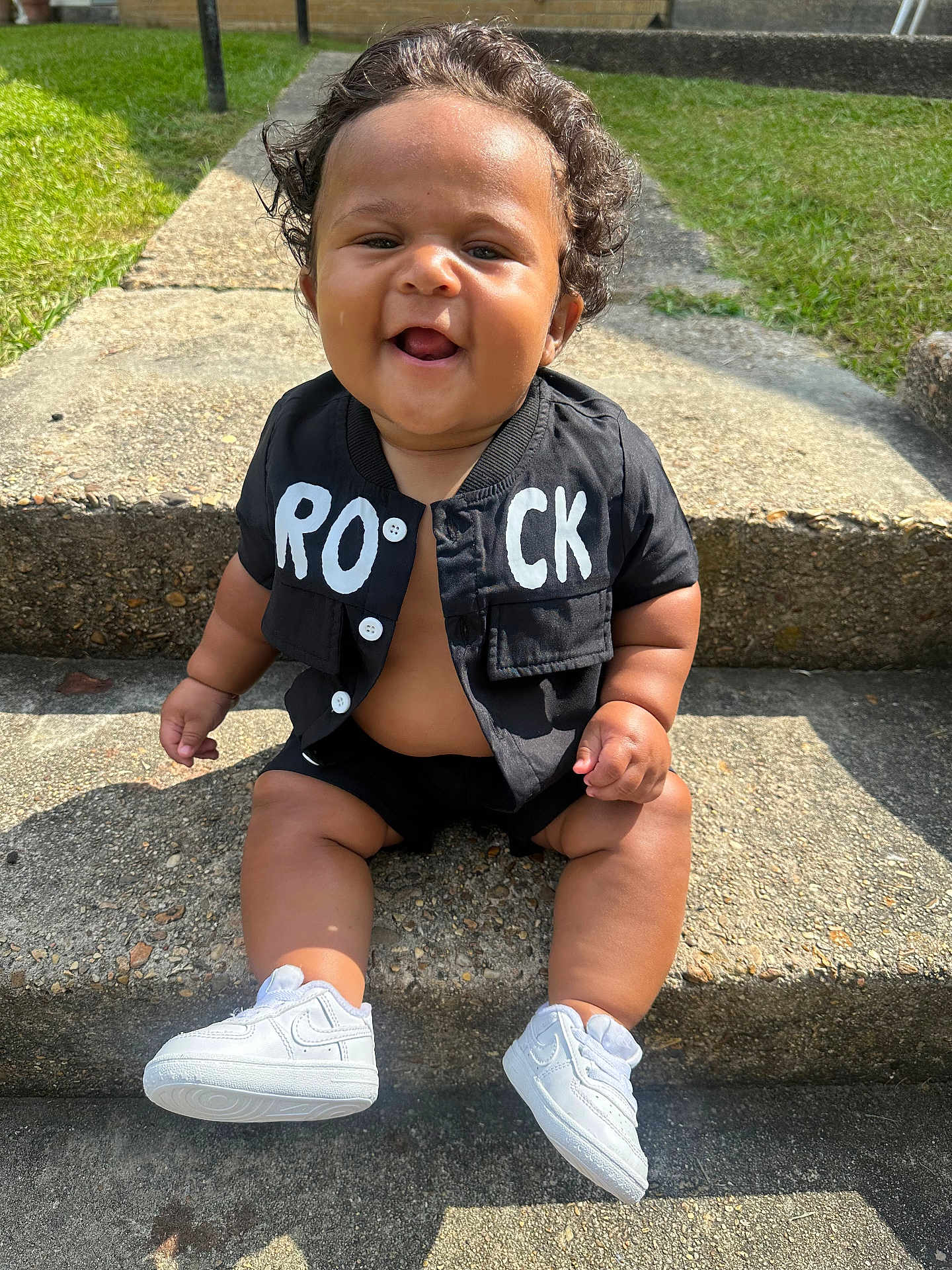 Zaelyn joined the competition — help win amazing prizes! baby, child, smiling, curly_hair, sitting, outdoor, steps, concrete, black_jacket, white_sneakers, happy, cute, sunlight, grass, casual_clothing, portrait, person, infant, small_shoes, daytime