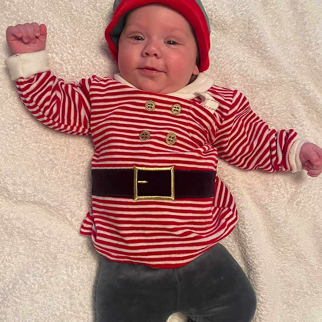 Marley is registered to the contest to win money with this photo: baby, bare_feet, blanket, child, cozy, cute, festive, grey_pants, hat, holiday_outfit, indoors, infant, lying_down, portrait, red, smile, soft_texture, striped_clothing, warm_clothing, white