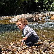 Maël a rejoint le concours — aidez-le/la à gagner de superbes lots ! child, toddler, curly_hair, river, rocks, water, outdoor, nature, squatting, sunlight, stream, pebbles, exploring, playful, daytime, casual_clothing, greenery, small_person, riverbank, curiosity