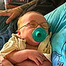 baby, blue_shirt, cartoon_print, casual_clothing, closeup, cozy, cute, glasses, hand, holding, indoor, infant, newborn, pacifier, person, portrait, relaxing, skin, sleepy, tattoo