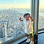 child, toddler, window, cityscape, skyline, sunset, building, urban, glass, floor, white_shirt, green_shorts, sneakers, smile, happy, indoors, reflection, view, standing, person