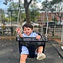 black_swing, blue_clothing, blurred_background, car, child, daytime, feet, fence, fun, happy, holding, outdoor, park, playground, seat, shoe, smiling, swing, toddler, tree