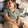 bed, beige_pants, casual_clothing, child, cozy, cute, expression, face, hair, home, indoor, person, playful, polo_shirt, portrait, relaxing, sitting, smiling, toddler, young_child