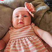 Maeve is registered to the contest to win money with this photo: arm, baby, blanket, cheeks, child, clothing, cute, face, flower_headband, headwear, indoors, infant, laying, pink_headband, plush, portrait, smile, soft_texture, striped_dress, young_child