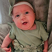 Maeve is registered to the contest to win money with this photo: baby, infant, green_dress, headband, flower_decoration, blanket, soft_texture, cute, portrait, child, young, smiling, face, eyes, hand, clothing, lying_down, indoor, closeup, adorable