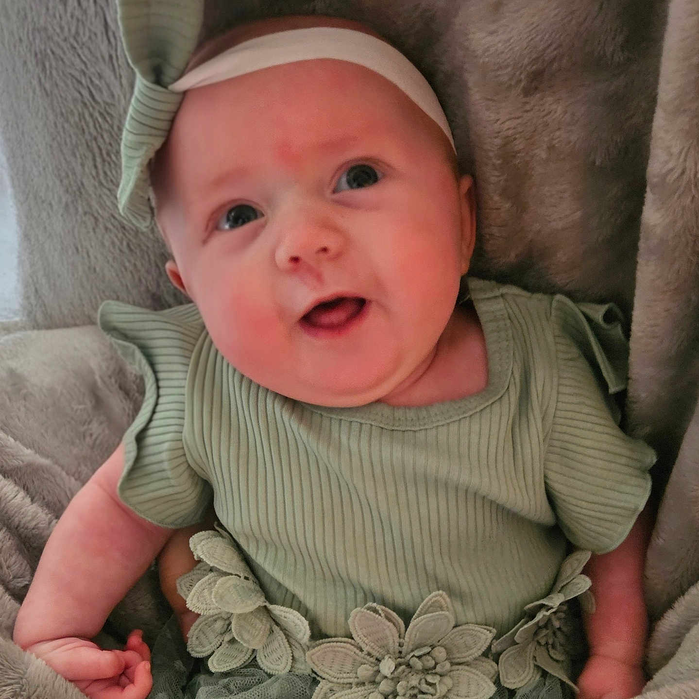 Maeve is registered to the contest to win money with this photo: adorable, baby, blanket, child, closeup, clothing, cute, eyes, face, flower_decoration, green_dress, hand, headband, indoor, infant, lying_down, portrait, smiling, soft_texture, young