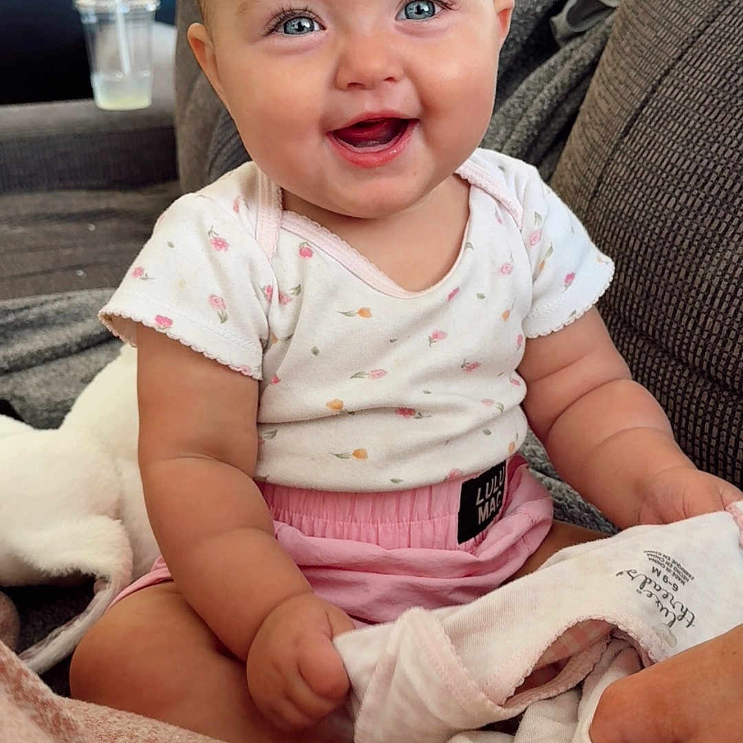 Sutton joined the competition — help win amazing prizes! baby, child, infant, smiling, happy, portrait, sitting, indoor, home, couch, blanket, clothing, pink, floral, blue_eyes, face, kitchen, bottle, cup, photography