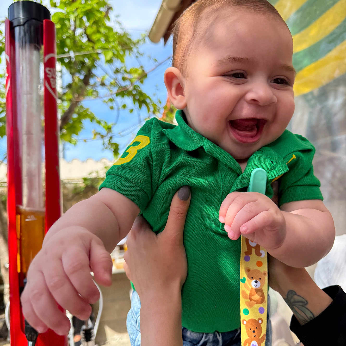 Guilherme participe au concours pour gagner de l'argent avec cette photo : baby, child, smiling, green_shirt, denim_shorts, happy, outdoor, daylight, tree, sky, hand, tattoo, person, face, clothing, playful, laughter, striped_awning, strap, cute