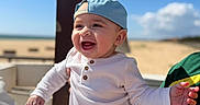 Guilherme participe au concours pour gagner de l'argent avec cette photo : baby, child, smiling, happy, outdoor, sunny, blue_sky, cap, striped_pants, person, portrait, holding, laughing, clothing, hat, cute, summer, beach, daylight, casual