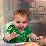 Guilherme participe au concours pour gagner de l'argent avec cette photo : baby, child, green_shirt, smiling, happy, highchair, hand, adult, watch, table, indoor, blurred_background, person, cafe, expression, cute, young_child, short_hair, playful, interaction