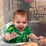 baby, child, green_shirt, smiling, happy, highchair, hand, adult, watch, table, indoor, blurred_background, person, cafe, expression, cute, young_child, short_hair, playful, interaction