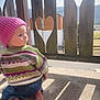 child, toddler, baby, pink_beanie, knit_hat, striped_sweater, jeans, bare_feet, wooden_fence, heart_cutout, balcony, outdoors, sunlight, shadow, portrait, side_profile, candid, countryside, railing, concrete_floor