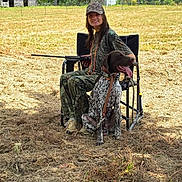 Moses is registered to the contest to win money with this photo: barn, camouflage_clothing, canine, chair, dog, field, grass, happy, hunting, leash, nature, outdoor, person, pet, rifle, sitting, sky, smiling, sunny, trees