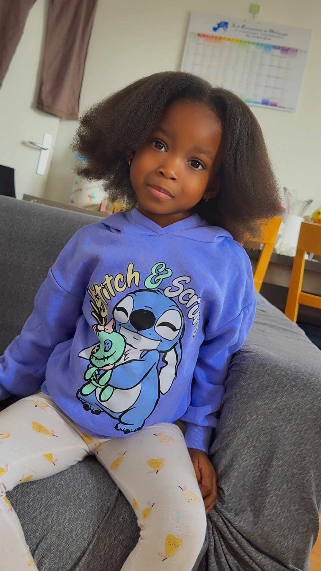 Aminata participe au concours pour gagner de l'argent avec cette photo : child, girl, hoodie, stitch, scrump, sitting, couch, indoor, hair, curly_hair, clothing, pants, patterned_pants, person, face, portrait, furniture, calendar, chair, casual